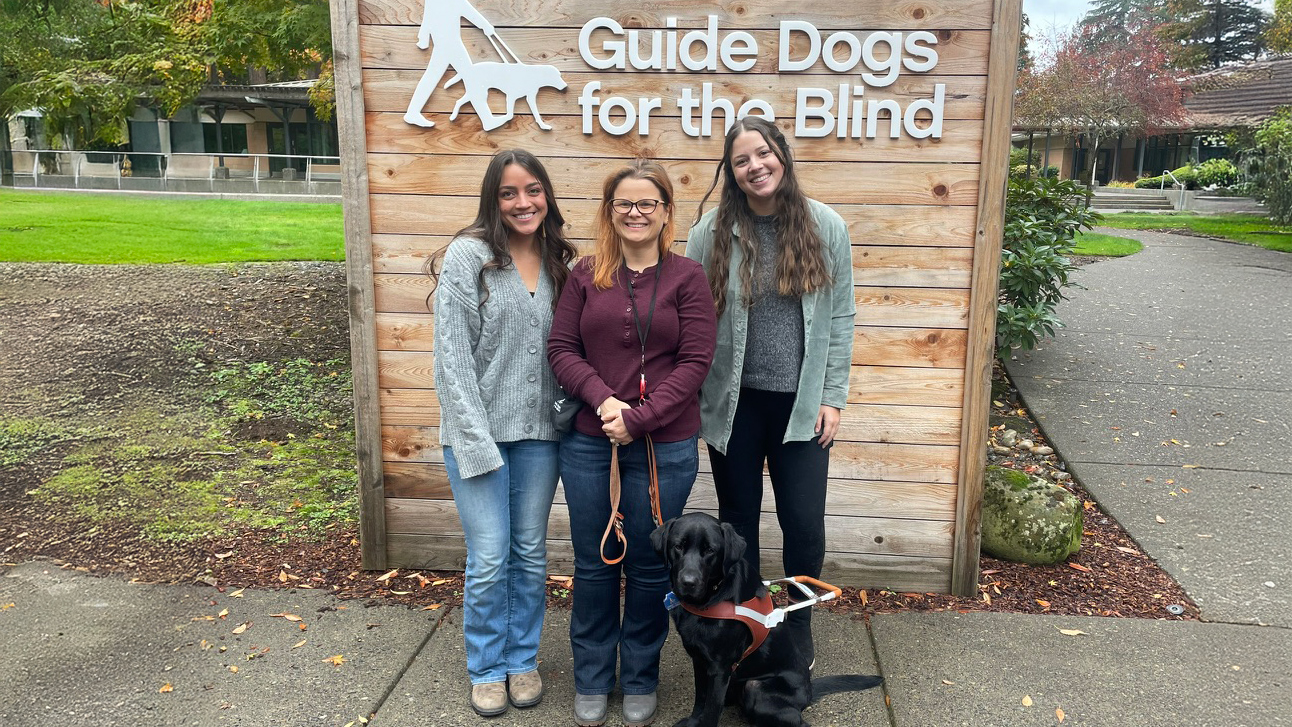 Three women and one black lab dog in front of sign that says Guide Dogs for the Blind