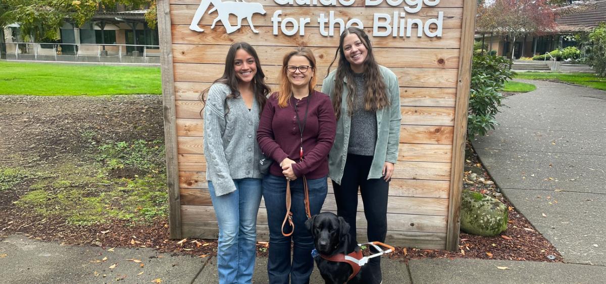 Three women and one black lab dog in front of sign that says Guide Dogs for the Blind