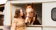 Horse looks out a horse trailer window. Woman stands next to it.  