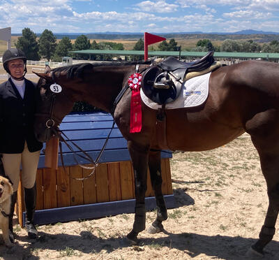 Woman stands next to horse sporting aribbon from an equine event. Dog stands with them on leash.