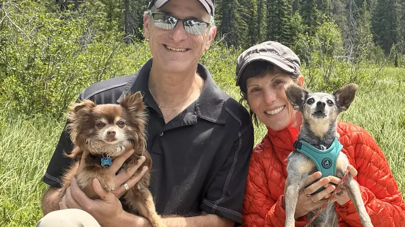 Two people each holding a dog on a sunny day in front of a forest