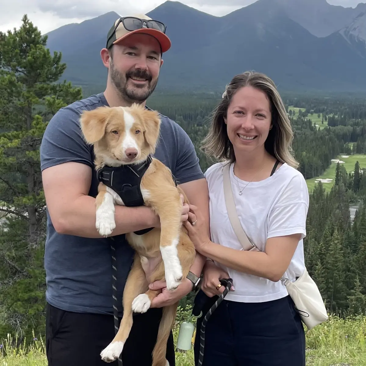 Man holds dog and stands next to woman. Mountain and river in background.
