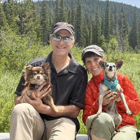 Two people each holding a dog on a sunny day in front of a forest