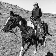 Black and white photo of a horse walking across grasslands with their owner sitting on their back 