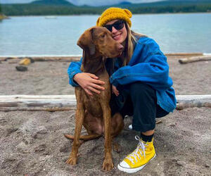 Smiling woman hugs dog on beach by a lake. 