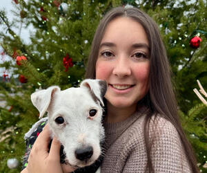 Woman holds small white dog while standing in front of Christmas tree.