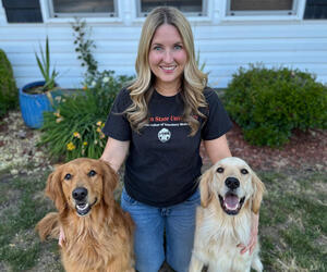 Woman kneeling outside with arms around two dogs. 